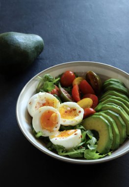 Akdeniz salatası, avokado ile haşlanmış yumurta, kremalı yumurta ve roket yatağında vişneli domates, EVO yağı, tuz ve limon suyu. Tek ve dengeli yemek. koyu arkaplan.