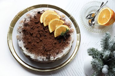 Lezzetli çikolata ve portakallı tiramisu keki, kakao ve çikolata parçalarıyla tozlanmış, portakal dilimleri ve biberiye ile süslenmiş, altın tepsiye konmuş beyaz bir tabakta servis ediliyor. Noel süsü. İtalyan tatlısı.