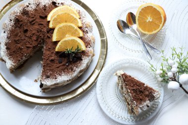 Lezzetli çikolata ve portakallı tiramisu keki, kakao ve çikolata parçalarıyla tozlanmış, portakal dilimleri ve biberiye ile süslenmiş, altın tepsiye konmuş beyaz bir tabakta servis ediliyor. Noel süsü. İtalyan tatlısı.