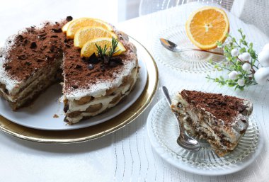 Lezzetli çikolata ve portakallı tiramisu keki, kakao ve çikolata parçalarıyla tozlanmış, portakal dilimleri ve biberiye ile süslenmiş, altın tepsiye konmuş beyaz bir tabakta servis ediliyor. Noel süsü. İtalyan tatlısı.