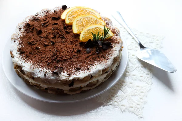 Çikolata ve portakallı tiramisu keki, kakao ve çikolata parçalarıyla tozlanmış, portakal dilimleri ve biberiye ile süslenmiş, beyaz arka planda izole edilmiş beyaz bir tabakta servis ediliyor. İtalyan tatlısı. Genel Görünüm.