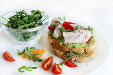 Fırında ekmek, avokado kremalı bruschetta, pişmiş jambon ya da hindi, ikiye bölünmüş kiraz domatesli, zeytinyağlı, taze roketli ve mandalinli ya da portakallı. Beyaz arkaplan.