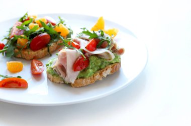 İki avokado tostlu tabak. İki parça tost da avokado kremalı. Bir bruschetta taze roka, domates ve portakal dilimleriyle kaplıdır. Diğeri pişmiş jambon, hindi, kiraz domatesleri ve roketle tatlandırılmış..