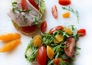 İki avokado tostlu tabak. İki parça tost da avokado kremalı. Bir bruschetta taze roka, domates ve portakal dilimleriyle kaplıdır. Diğeri pişmiş jambon, hindi, kiraz domatesleri ve roketle tatlandırılmış..