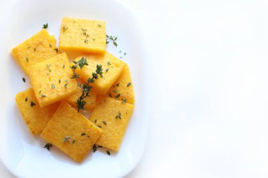 Bir tabak polenta ya da polenta cipsi, dilimlenmiş ve kızartılmış ya da pişirilmiş, taze kekik ile tatlandırılmış, beyaz arka plan. Mısır unu ve sudan yapılmış. Genel bakış. İtalyan yemeği..