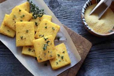Bir tabak polenta veya polenta cipsi, dilimlenmiş ve kızartılmış veya pişirilmiş, taze kekik ile tatlandırılmış, ahşap arka plan. Mısır unu ve sudan yapılmış. Genel bakış. İtalyan yemeği..