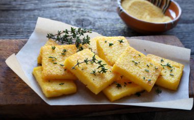Bir tabak polenta veya polenta cipsi, dilimlenmiş ve kızartılmış veya pişirilmiş, taze kekik ile tatlandırılmış, ahşap arka plan. Mısır unu ve sudan yapılmış. Genel bakış. İtalyan yemeği..