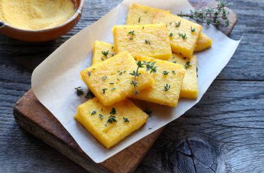Bir tabak polenta veya polenta cipsi, dilimlenmiş ve kızartılmış veya pişirilmiş, taze kekik ile tatlandırılmış, ahşap arka plan. Mısır unu ve sudan yapılmış. Genel bakış. İtalyan yemeği..