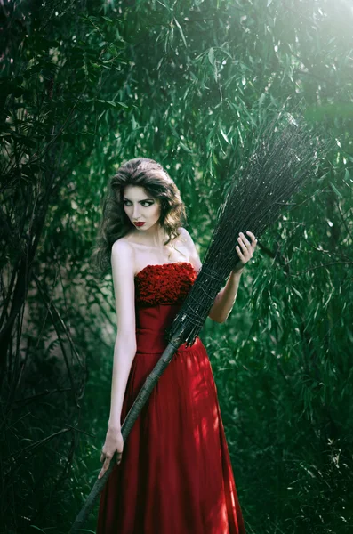 Beautiful girl witch standing in the woods with a broom and evil looks away — Stock Photo, Image