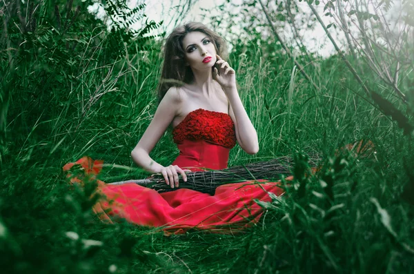 Hermosa chica, bruja sentada en la hierba en un vestido rojo sosteniendo su mano sobre su escoba, la otra mano mira su mejilla —  Fotos de Stock