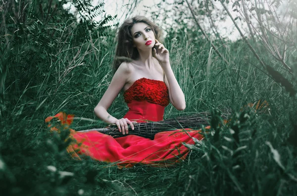 Красивая девушка, ведьма сидит на траве в красном платье держа руку на метле, другая рука смотрит на его щеку — стоковое фото