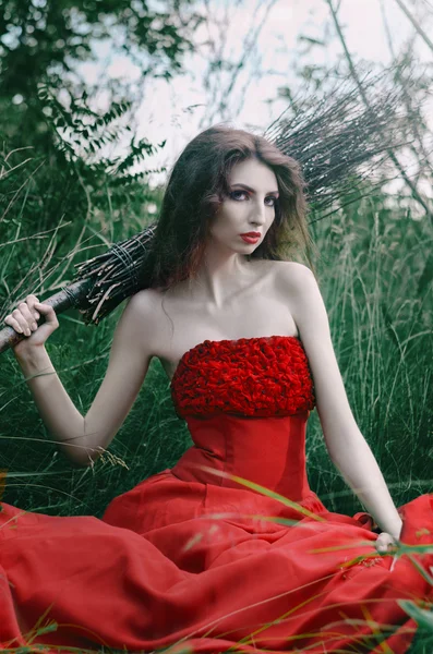 Belle fille, sorcière assise sur l'herbe dans une robe rouge avec un balai sur son épaule — Photo