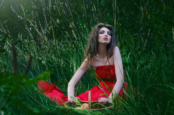 Hermosa bruja sentada en la hierba en el bosque y sosteniendo una cesta con manzanas —  Fotos de Stock