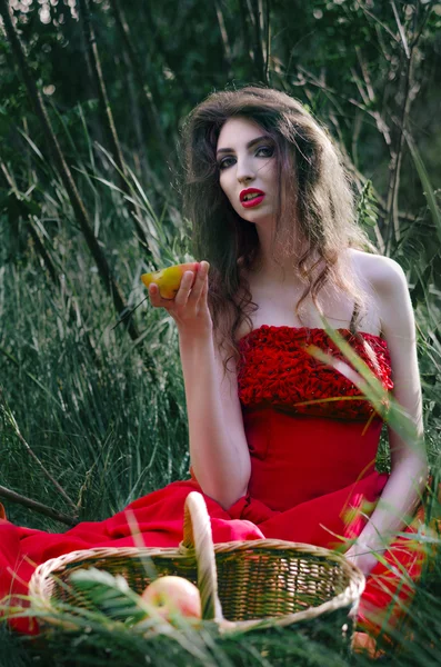 Hermosa chica, bruja sentada en la hierba en un vestido rojo y comer media manzana que lleva a la cesta —  Fotos de Stock