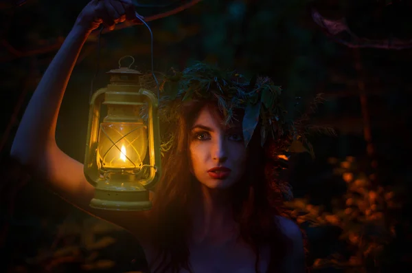 Красива дівчина Холдинг ліхтар з вінок на його голову стоїть осібно в лісі. Відьма висвітлює свій шлях. Хеллоуїн. — стокове фото