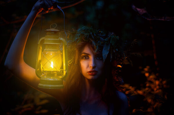 Beautiful Girl Holding a Lantern with a Wreath on his Head Stands Alone in the Woods. Witch Illuminates your way. Halloween.