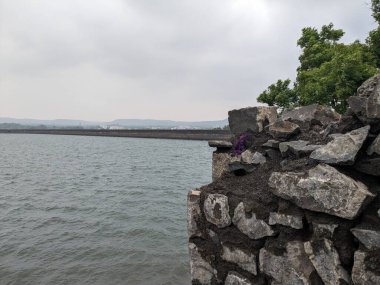 Engebeli bir taş duvar dalgalı suyla gri tonlu bir göle dalıyor, bulutlu gökyüzü tarafından çerçeveleniyor ve uzaktan belli belirsiz görünen tepeler.