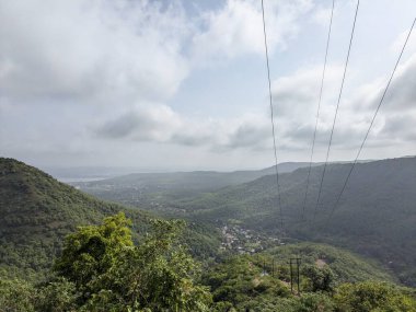 Geniş bir manzara ormanlık dağları yuvarlıyor ve aşağıdaki vadiye yerleşmiş küçük bir köy, yoğun ve bulutlu bir gökyüzünün altında ön planda uzanan önemli elektrik hatları bulunuyor..