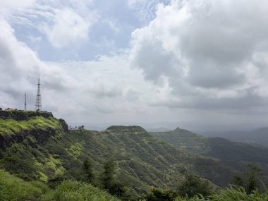 Yüksek bir dağ manzarasından nefes kesici panoramik bir manzara yemyeşil orman tepelerinin katmanlarını ve yumuşak, bulutlu bir gökyüzünün altındaki vadide yuva yapmış uzak bir yerleşim yerini ortaya çıkarıyor..