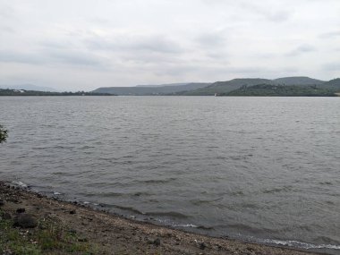 Göl üzerinde bulutlu bir gökyüzü, taştan bir sahile vuran hafif dalgalarla tepelik arazi ve su üzerinde görünen beyaz binalar..