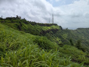 Yüksek bir dağ manzarasından nefes kesici panoramik bir manzara yemyeşil orman tepelerinin katmanlarını ve yumuşak, bulutlu bir gökyüzünün altındaki vadide yuva yapmış uzak bir yerleşim yerini ortaya çıkarıyor..