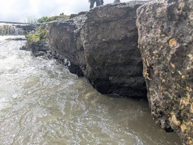 Çalkantılı nehir suyunun geniş ve yıpranmış bir kayanın yanından akışının detaylı fotoğrafları. Görüntü, taşın dokusunu ve bulanık suyun dinamik hareketini yakalar..