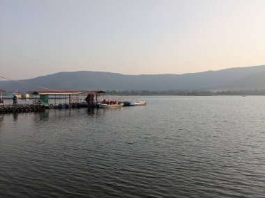 Huzurlu göl manzarası, tepeli bir iskeleye yanaşmış tekneleri ve mesafeye uzanan açık suları gösteriyor.