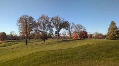 Şehir parkındaki büyük bir golf sahasının manzarası. Üzerinde yapraklı ağaçlar ve mavi gökyüzü var.
