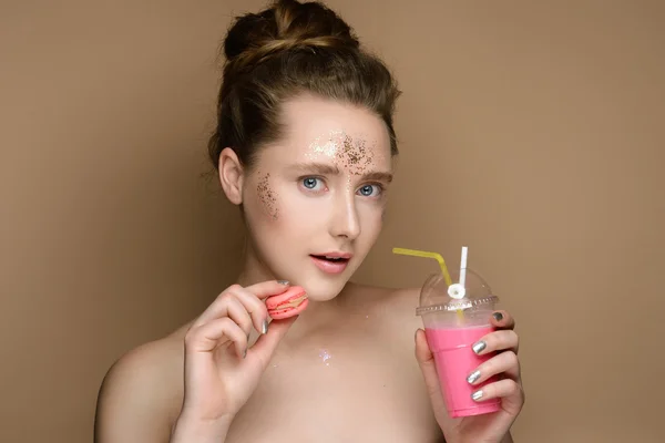 Portrait de jeune belle fille avec coctail shake au lait rose et macaron dans les mains sur le fond doré. Maquillage nu avec tinsel et updo. Concept de la beauté . — Photo