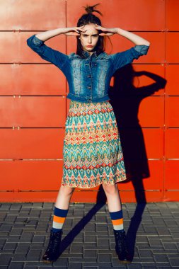 Young teenager girl having fun, posing and smiling near red wall background in skirt and jeans jacket on the sunset.