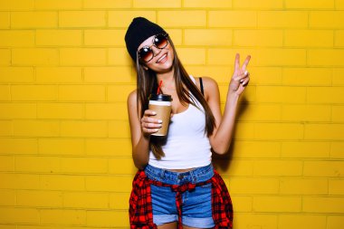 Beautiful young sexy girl drink coffee, smiling and posing near yellow wall background in sunglasses, red plaid shirt.