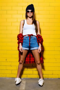 Beautiful young sexy girl posing and smiling near yellow wall background in sunglasses, red plaid shirt, shorts.