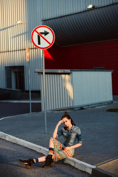 Young teenager girl sitting on the road outside near urban red wall background in skirt and jeans jacket on the sunset. — Stock Photo, Image