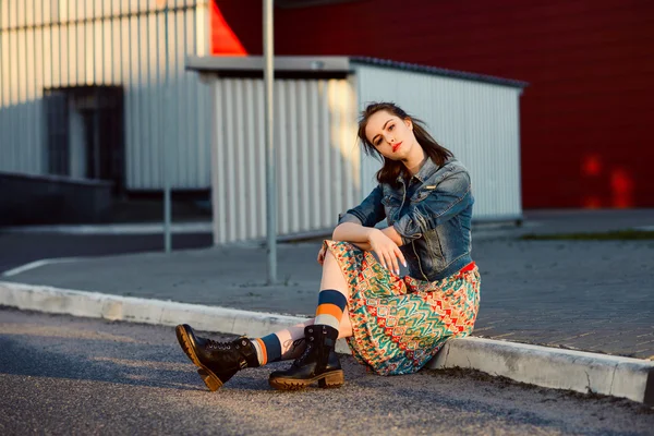Joven adolescente sentada en la carretera fuera cerca de fondo de pared roja urbana en falda y chaqueta jeans en la puesta de sol . —  Fotos de Stock