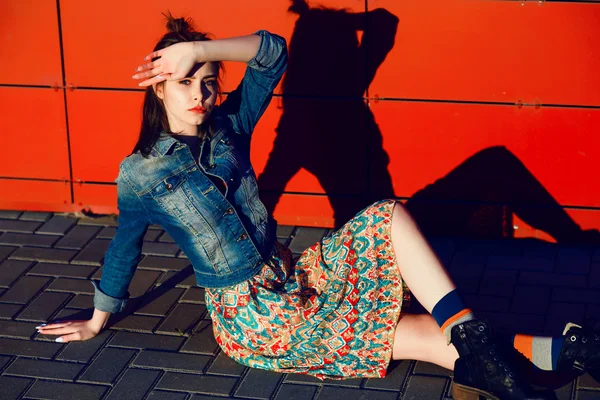 Young teenager girl posing and sitting near urban red wall background in skirt and jeans jacket on the sunset. — Stock Fotó