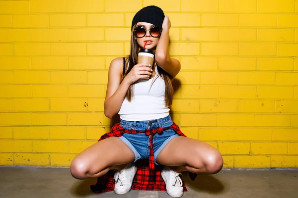 Hermosa chica sexy joven beber café y sentado cerca de fondo amarillo de la pared en gafas de sol, camisa de cuadros rojos, pantalones cortos . —  Fotos de Stock