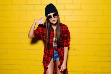 Güzel genç seksi hipster kız poz ve güneş gözlüğü kentsel sarı duvar arka plan yakınında gülümseyen, kırmızı ekose gömlek, şort, şapka.