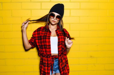 Beautiful young sexy hipster girl posing and smiling near urban yellow wall background in sunglasses, red plaid shirt, shorts, hat. clipart