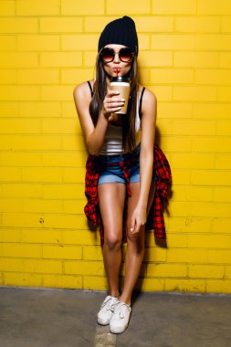 Güzel genç seksi hipster kız kahve içmek, gülümseyen ve güneş gözlüğü kentsel sarı duvar arka plan yakınında poz, kırmızı ekose gömlek, şort, şapka.