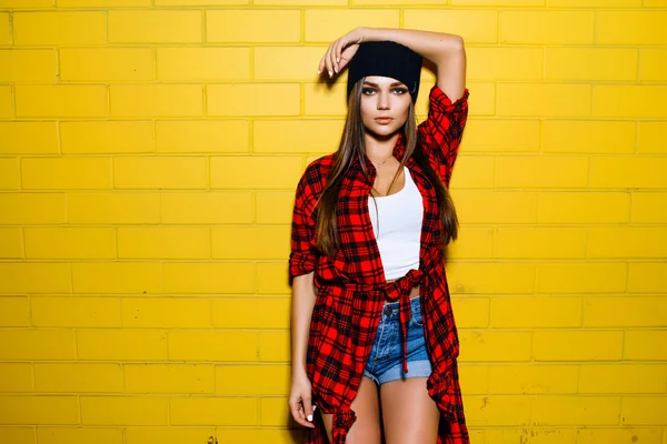 Hermosa chica hipster sexy joven posando y sonriendo cerca de fondo de pared amarilla urbana en camisa a cuadros rojo, pantalones cortos, sombrero . —  Fotos de Stock
