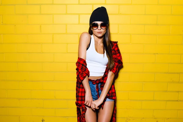 Hermosa chica hipster sexy joven posando y sonriendo cerca del fondo de la pared amarilla urbana en gafas de sol, camisa a cuadros roja, pantalones cortos, sombrero, zapatillas de deporte . —  Fotos de Stock