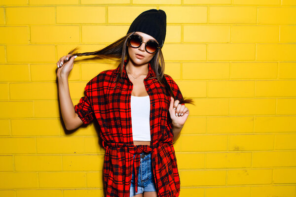 Beautiful young sexy hipster girl posing and smiling near urban yellow wall background in sunglasses, red plaid shirt, shorts, hat.