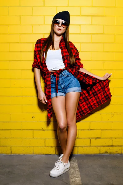Hermosa chica hipster sexy joven posando y sonriendo cerca del fondo de la pared amarilla urbana en gafas de sol, camisa a cuadros roja, pantalones cortos, sombrero, zapatillas de deporte . — Foto de Stock