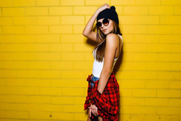 Красивая молодая сексуальная хипстерша позирует и улыбается на фоне городской желтой стены в солнечных очках, красной клетчатой рубашке, шортах, шляпе, кроссовках . — стоковое фото