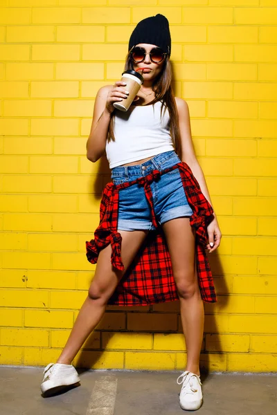 Bella giovane ragazza hipster sexy bere caffè, sorridente e posa vicino urbano giallo sfondo della parete in occhiali da sole, camicia scozzese rossa, pantaloncini, cappello . — Foto Stock