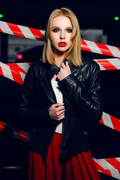 Mode portret van sexy blonde meisje met rode lippen dragen een rock zwarte stijl en rode broek poseren en glimlachend op de achtergrond van de waarschuwing tape. Zwart, rood, wit. — Stockfoto