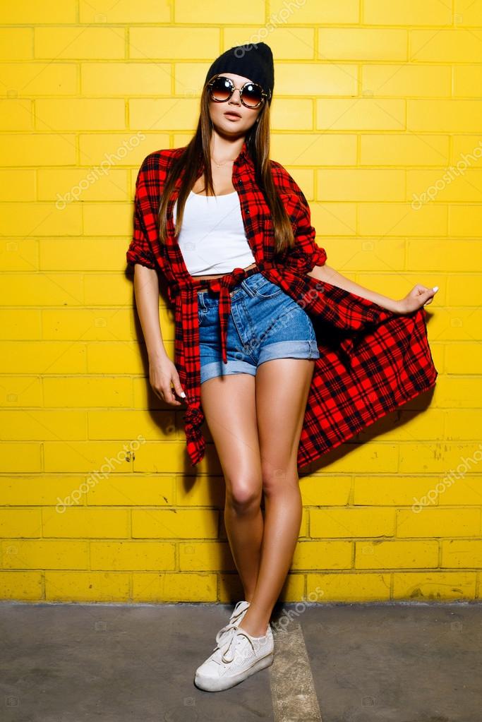 Beautiful young sexy hipster girl posing and smiling near urban yellow wall  background in sunglasses, red plaid shirt, shorts, hat, sneakers. Stock  Photo by ©isdvsk.gmail.com 111722462