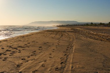 Gün batımında sakin sahil manzarası, kumlu sahilde yumuşak dalgalar, mesafeye doğru giden ayak izleri, ve barışçıl bir atmosfer yaratan dağlar.