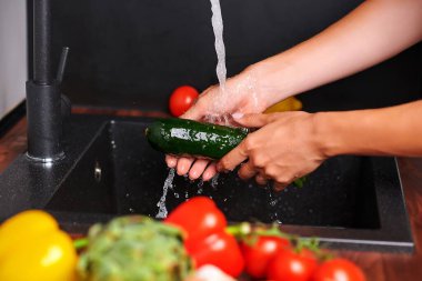 Bir çift el, domates ve biber gibi canlı sebzelerle çevrili şık bir mutfak lavabosunda akan suyun altında taze bir salatalığı yıkıyor.