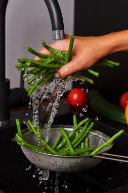 Bir el, akan suyun altında taze yeşil fasulyeleri yıkıyor, sebzelerin canlı renklerini ve çeşitli malzemelerle temiz, modern mutfak ortamını vurguluyor.